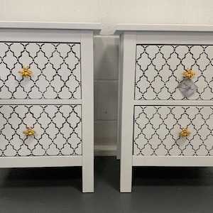 May include: Two white bedside tables with two drawers each. The drawers feature a black and white geometric pattern and gold bee-shaped knobs.