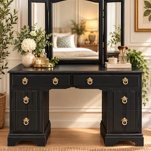 May include: A black vanity table featuring a tri-fold mirror, two wide top drawers, and four smaller side drawers. The furniture piece is accented with ornate gold-toned ring pulls on each drawer front and has a smooth, flat tabletop surface.