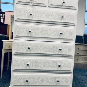 May include: White chest of drawers with six drawers. Each drawer has a silver knob and a floral stencil design.