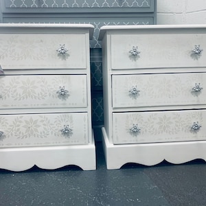 May include: Two white wooden bedside tables with three drawers each. The drawers feature a white floral design and silver bee-shaped knobs.