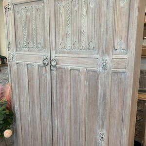May include: A whitewashed wooden cabinet with two doors and intricate carved details. The cabinet has a distressed finish and features ornate metal hinges.