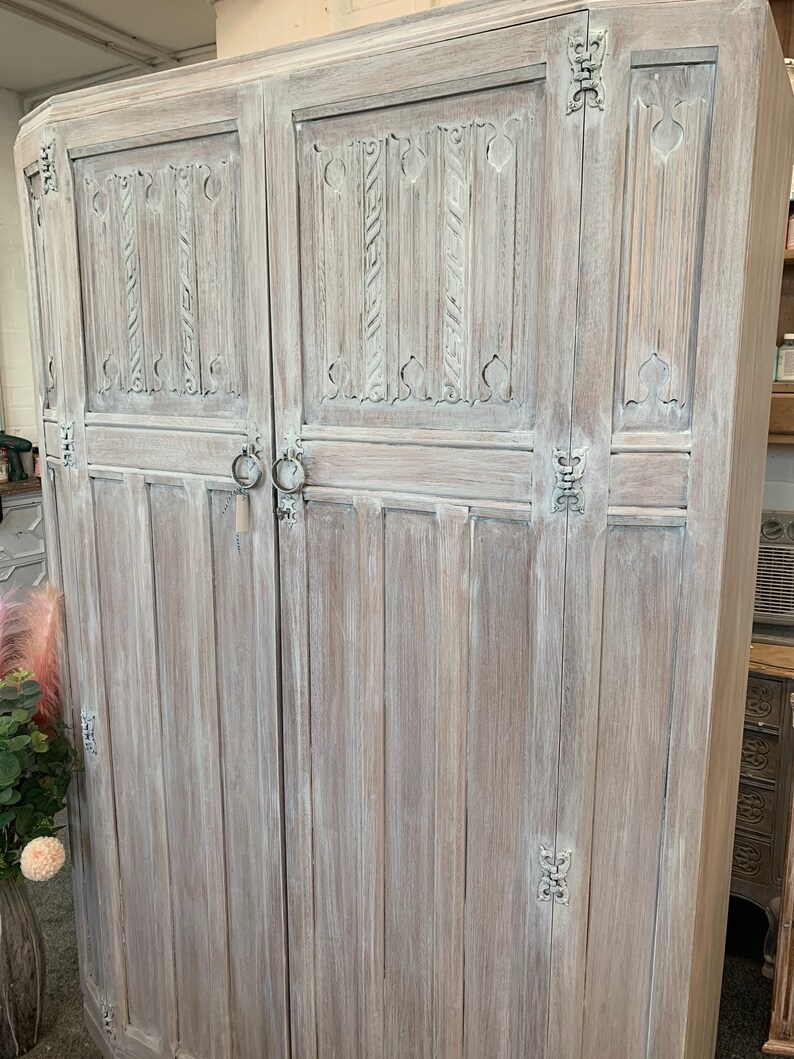 May include: A whitewashed wooden cabinet with two doors and intricate carved details. The cabinet has a distressed finish and features ornate metal hinges.