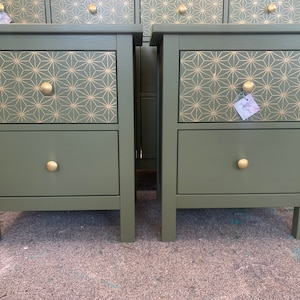 May include: Two sage green bedside tables with two drawers each. The top drawer has a gold geometric pattern. Each drawer has a gold knob. The tables are made of wood and have a simple, modern design.