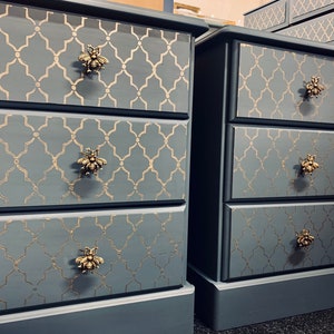 May include: Two grey bedside tables with three drawers each. The drawers have gold bee-shaped knobs and a gold geometric pattern.