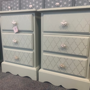 May include: A pair of light blue bedside cabinets, each with three drawers. The drawers are decorated with a repeating grey pattern and silver-toned knobs. The cabinets have a scalloped base and a decorative top. (Approx. dimensions in cm)