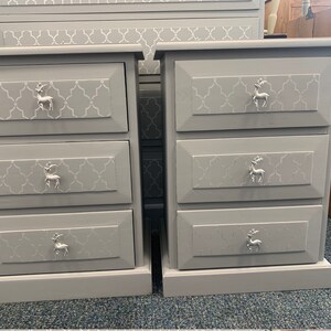 May include: Two grey wooden bedside tables with three drawers each. The drawers have a white geometric pattern and silver deer-shaped knobs.