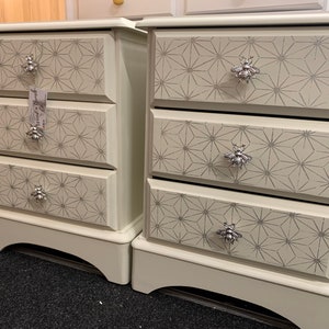May include: Two white bedside tables with three drawers each. The drawers have a geometric pattern and bee-shaped handles.