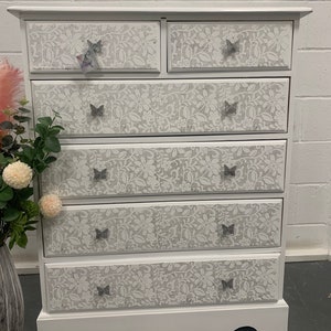 May include: White chest of drawers with five drawers. Each drawer has a floral pattern and a butterfly-shaped knob.