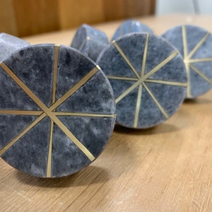 May include: A collection of grey marble cabinet knobs featuring a gold starburst pattern. The knobs are circular and have a polished finish. They are displayed on a light wooden surface.
