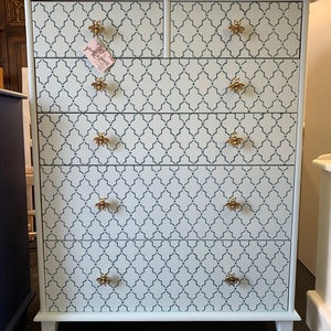 May include: A white six-drawer chest of drawers with a geometric pattern in a darker blue. The chest has gold-coloured bee-shaped handles. The chest is a light blue colour and has a tag that says "Wood & Brush".