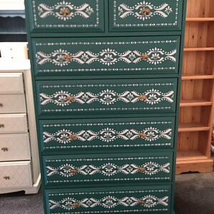 May include: A dark teal chest of drawers with seven large drawers and two smaller top drawers. It features white floral stencilled patterns and gold-coloured handles. A piece of furniture.