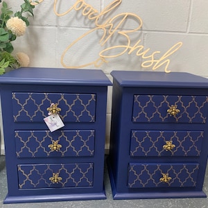 May include: A pair of navy blue bedside tables, each with three drawers. The drawer fronts have a gold geometric design and bee-shaped handles. The tables are set against a white brick wall with a wooden sign that says "Wood Brush".