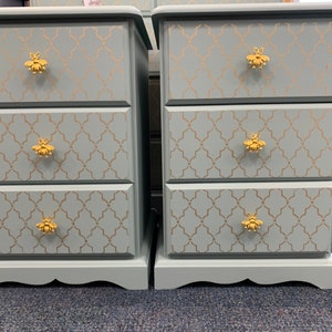 May include: Two light blue bedside tables with three drawers each. The drawers have gold bee-shaped knobs and a gold geometric pattern.