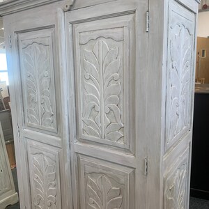 May include: A white painted wooden cabinet with two doors and carved leaf designs. The cabinet has a distressed finish and is a vintage style.