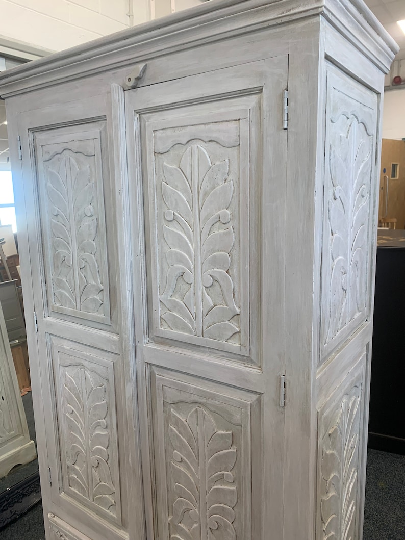 May include: A white painted wooden cabinet with two doors and carved leaf designs. The cabinet has a distressed finish and is a vintage style.