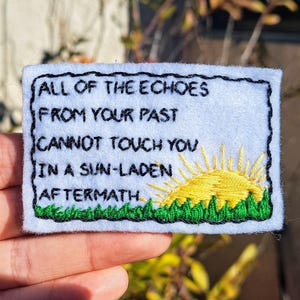 Puede incluir: Un parche de fieltro blanco con bordado negro que dice "All of the echoes from your past cannot touch you in a sun-laden aftermath". El bordado incluye un sol amarillo que sale sobre hierba verde.