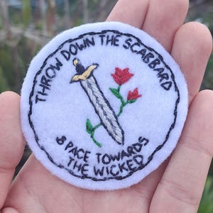 May include: White embroidered patch with a black outline. The patch features a sword with a yellow handle, a red rose, and the text "Throw down the scabbard & pace towards the wicked".