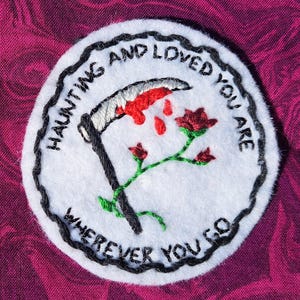 May include: White embroidered patch with black stitching. The patch features a black scythe with red dripping blood, a red rose, and the text "Haunting and loved you are wherever you go."