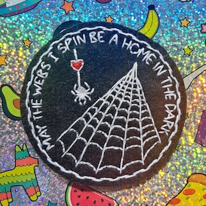 May include: A round, black embroidered patch with white stitching. The patch features a spider web design and the text "MAY THE WEBS I SPIN BE A HOME IN THE DARK". A spider and a heart are also embroidered on the patch.