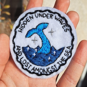 May include: A white embroidered patch with a blue whale tail and the text "Hidden Under Waves And Lost Amongst The Sea".