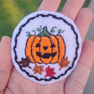 May include: A white felt patch with a black stitched border, featuring an embroidered orange pumpkin with a jack-o'-lantern face and green stem.  The pumpkin is surrounded by brown and orange embroidered leaves.