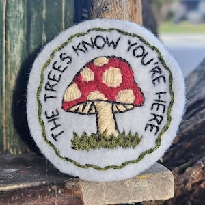 Pode incluir: Patch bordado com um design de cogumelo vermelho com pontos brancos e um caule bege. O texto "THE TREES KNOW YOU'RE HERE" está em preto. Fundo branco e borda verde.