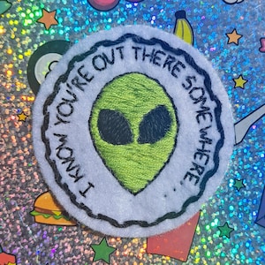 Puede incluir: Un parche redondo blanco con la frase bordada "I KNOW YOU'RE OUT THERE SOMEWHERE." El parche presenta una cabeza de alienígena verde con ojos negros en el centro, rodeada por un borde cosido negro. El fondo es una superficie holográfica y brillante.