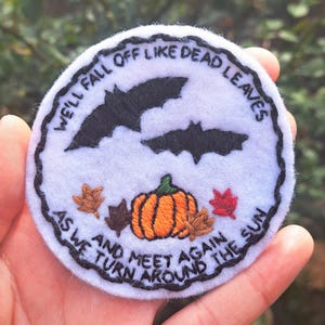 Może przedstawiać: Biała łatka z filcu z czarnymi nietoperzami, pomarańczową dynią i brązowymi liśćmi. Tekst brzmi "We'll fall off like dead leaves as we turn around the sun and meet again".