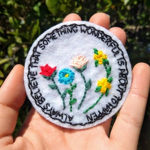 Puede incluir: Un círculo de fieltro blanco con bordado negro alrededor del borde. El texto "Siempre cree que algo maravilloso está a punto de suceder" está bordado en negro. Hay cinco flores bordadas en el centro: una flor roja, una flor azul, una flor rosa y dos flores amarillas.