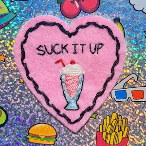 May include: Pink felt heart-shaped patch with black stitching around the edges. The patch has the text "SUCK IT UP" embroidered in black thread. A small embroidered milkshake with a cherry on top is below the text.