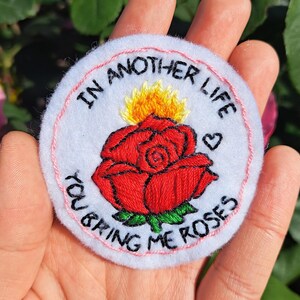 Può includere: Toppa ricamata con una rosa rossa, foglie verdi e una fiamma gialla, circondata dal testo "IN ANOTHER LIFE YOU BRING ME ROSES". La toppa ha uno sfondo bianco, un bordo rosa e un piccolo cuore.