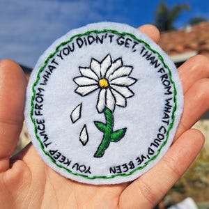 May include: White embroidered patch with a green daisy and the text "You keep twice from what you didn't get, than from what could've been."