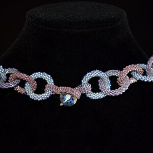 May include: A beaded necklace with a chain-link design in shades of blue and purple. The necklace features a large, faceted, blue crystal pendant.