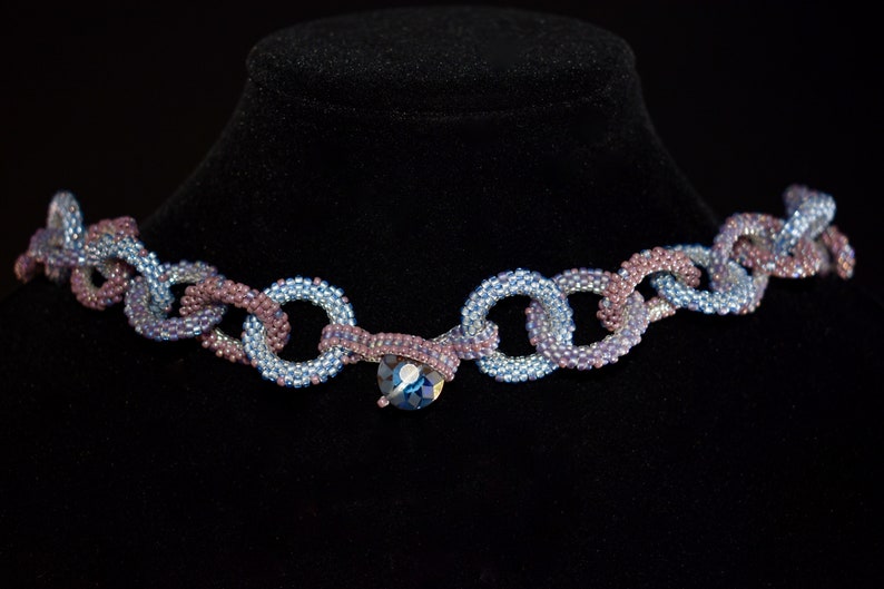 May include: A beaded necklace with a chain-link design in shades of blue and purple. The necklace features a large, faceted, blue crystal pendant.
