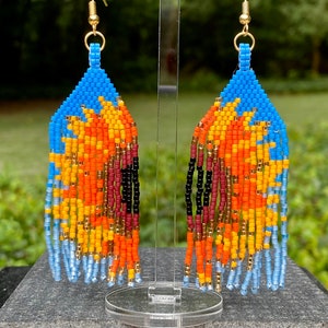 May include: A pair of beaded earrings with a blue, orange, yellow, red, and black color scheme. The earrings are hanging from gold hooks.