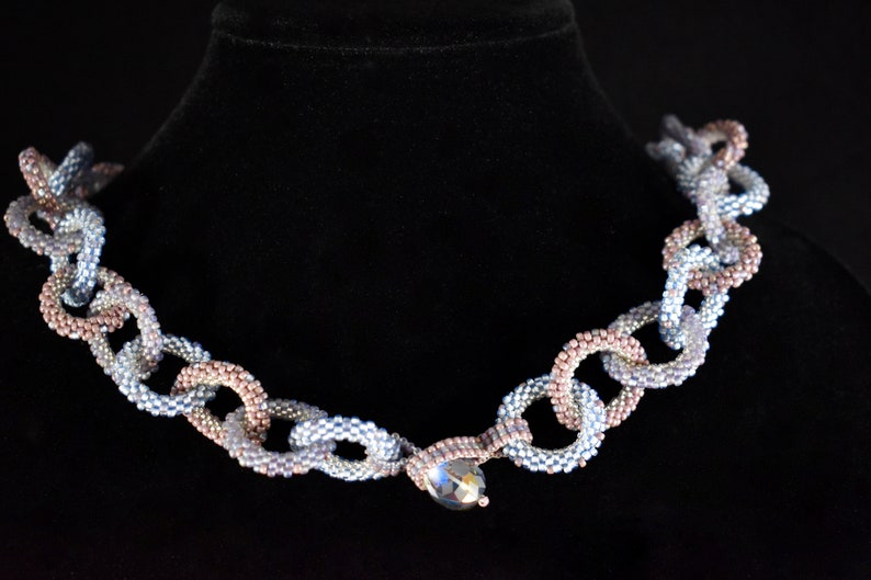 May include: A silver and rose gold beaded necklace with a large crystal pendant. The necklace is made of small, round beads that are strung together in a chain-like pattern.