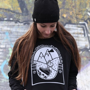 K&ouml;nnte beinhalten: Ein schwarzer, kurzer Kapuzenpullover mit einem wei&szlig;en Aufdruck eines Rollschuhs vor Bergen. Der Aufdruck enth&auml;lt auch den Text "Roll. Grow. Empower." Das Model tr&auml;gt eine schwarze Strickm&uuml;tze mit Schleife.