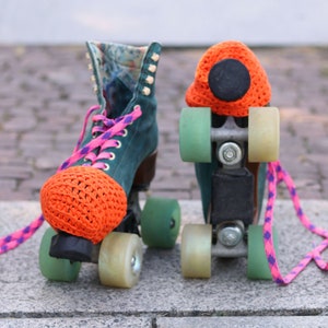 May include: A pair of vintage roller skates with orange crocheted covers on the wheels. The skates have green wheels and black frames. The laces are pink and purple.