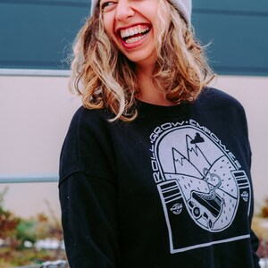 K&ouml;nnte beinhalten: Eine Frau tr&auml;gt eine graue M&uuml;tze mit dem Wort "MSCH" darauf, ein schwarzes Sweatshirt mit einer wei&szlig;en Grafik einer Bergkette und dem Text "Roll. Grow. Empower." und einem wei&szlig;en Logo unten rechts, lacht.