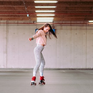 Peut inclure: Une personne portant un pull rose et un pantalon ray&eacute; fait du roller dans un parking en b&eacute;ton. La personne porte des rollers rouges avec des roues noires.