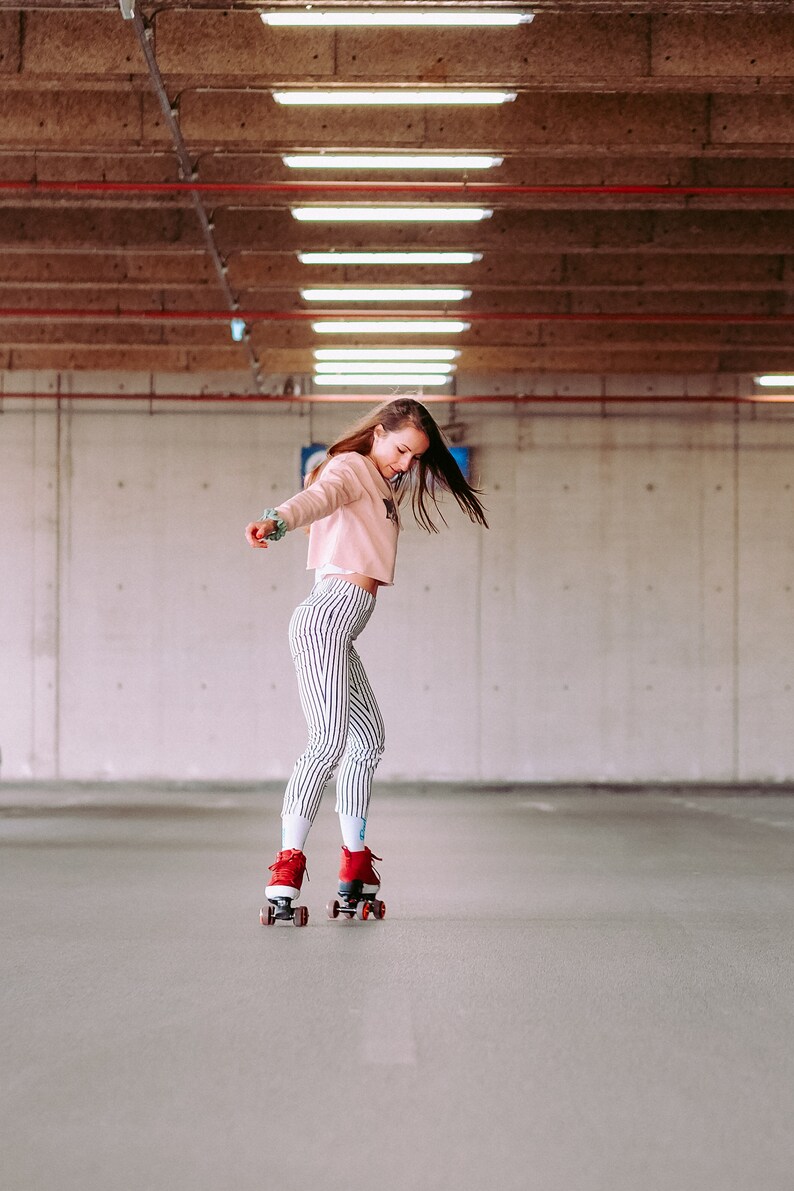 Peut inclure: Une personne portant un pull rose et un pantalon ray&eacute; fait du roller dans un parking en b&eacute;ton. La personne porte des rollers rouges avec des roues noires.