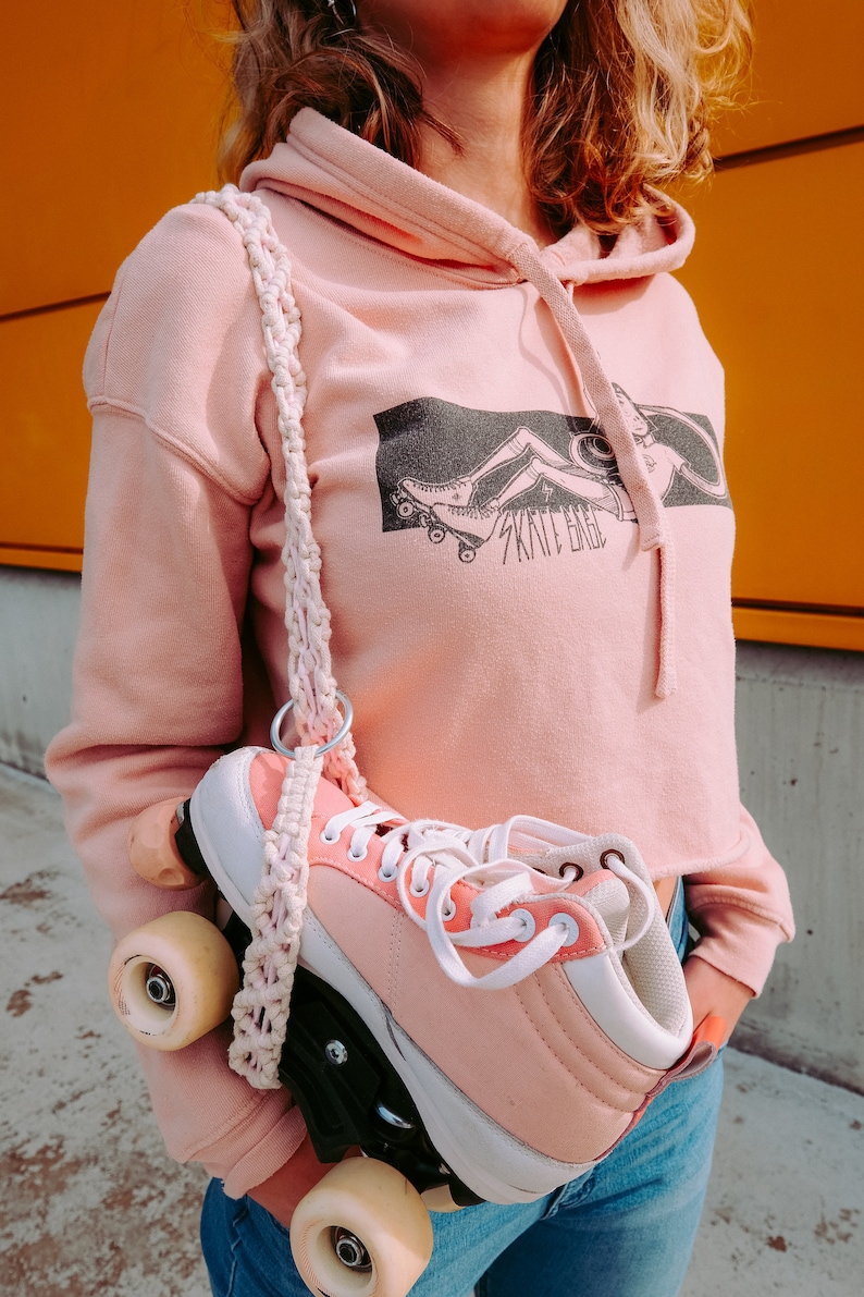 Peut inclure: Un sweat &agrave; capuche rose court avec un graphique d'une personne faisant du roller et le texte "Skate Babe". Le sweat est port&eacute; par une personne tenant une paire de rollers roses avec des lacets blancs.