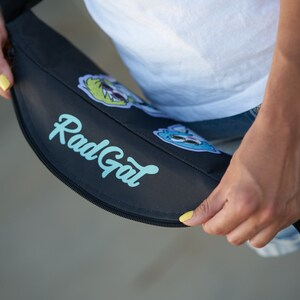 May include: A black fanny pack with a zipper closure and the text "Rad Gal" printed in light blue. The fanny pack has colorful cartoon patches.