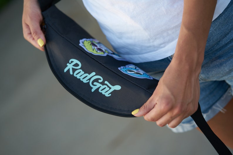 May include: A black fanny pack with a zipper closure and the text "Rad Gal" printed in light blue. The fanny pack has colorful cartoon patches.