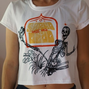May include: White cropped t-shirt with a graphic print of a skeleton holding a sign that says "Stay Weird until we're Dead".