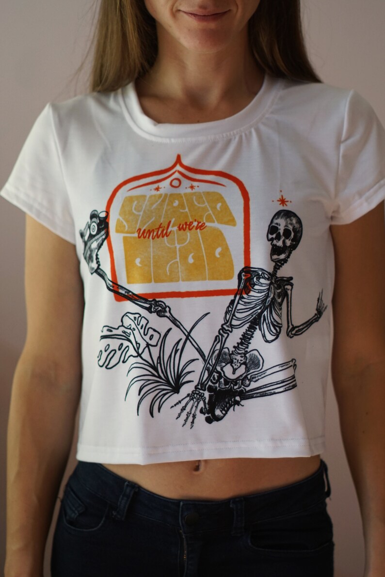 May include: White cropped t-shirt with a graphic print of a skeleton holding a sign that says "Stay Weird until we're Dead".