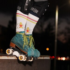 Op de afbeelding: Turquoise roller skates met witte wielen en groene veters. De skates hebben een wit-gele gestreepte sok met een bliksem-graphic.