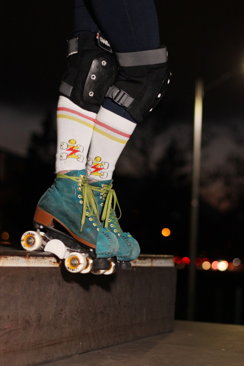 Op de afbeelding: Turquoise roller skates met witte wielen en groene veters. De skates hebben een wit-gele gestreepte sok met een bliksem-graphic.
