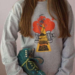 May include: A gray sweatshirt with a graphic of a skeleton skateboarding in a red cloud. The text "STAY IN THE WERE" is printed on the yellow ramp. A teal roller skate is being held in front of the sweatshirt.