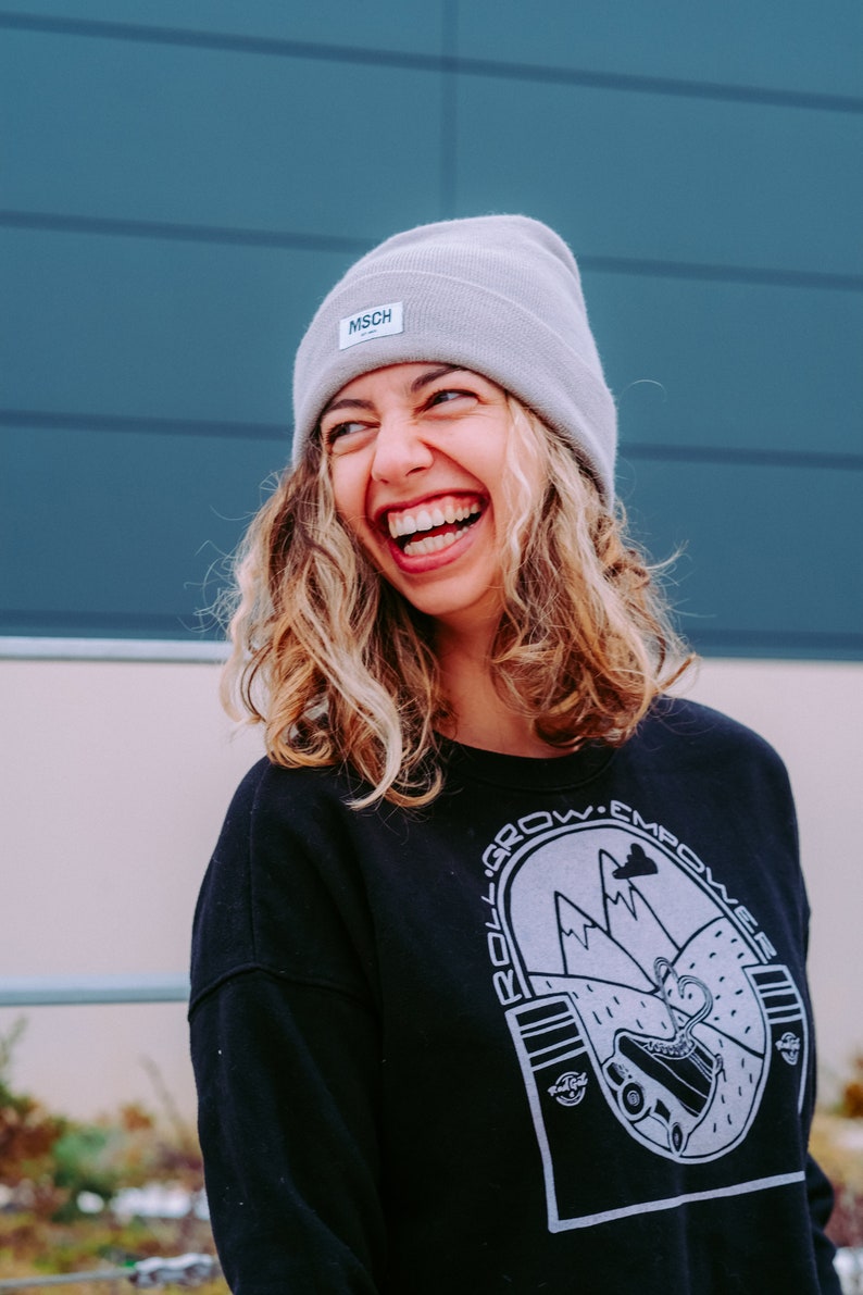 K&ouml;nnte beinhalten: Eine Frau tr&auml;gt eine graue M&uuml;tze mit dem Wort "MSCH" darauf, ein schwarzes Sweatshirt mit einer wei&szlig;en Grafik einer Bergkette und dem Text "Roll. Grow. Empower." und einem wei&szlig;en Logo unten rechts, lacht.